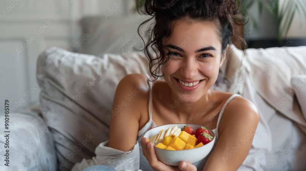 A young woman, with a radiant smile, holds a colorful bowl of fresh fruits while sitting comfortably on a cozy couch. A young woman, with a radiant smile, holds a colorful bowl of fresh fruits while sitting comfortably on a cozy couch.