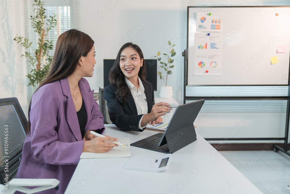 Two Asian businesswomen cheerfully discussing plans in office using calculator and laptop, standing in front of a whiteboard talking. Two Asian businesswomen cheerfully discussing plans in office using calculator and laptop, standing in front of a whiteboard talking.