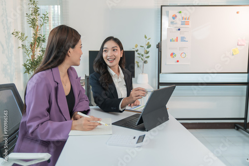 Two Asian businesswomen cheerfully discussing plans in office using calculator and laptop, standing in front of a whiteboard talking.