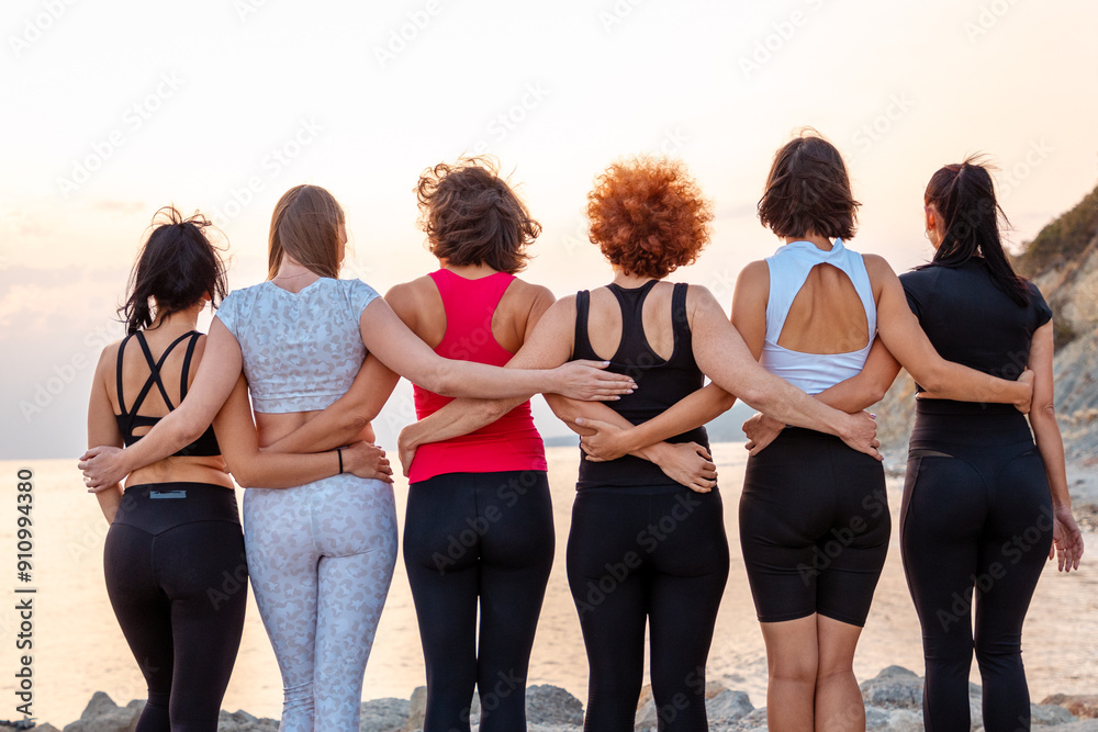 Fototapeta premium Rear view of group of fit women in sportswear stands in row and hugs each other. Light sunset sky in background. Copy space. Concept of girl power and sports competition