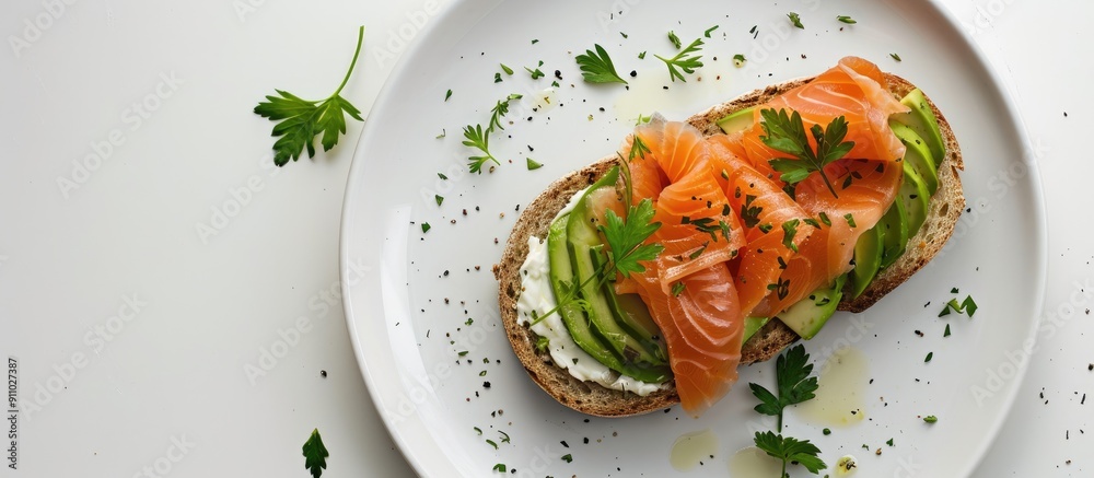 Top view of a healthy breakfast plate featuring a sandwich filled with smoked salmon, avocado, cream cheese, and parsley on white bread. The plate is set against a white background with ample copy