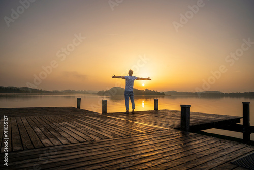 Carefree man with arms outstretched standing on pier near Woerthsee lake at sunrise photo