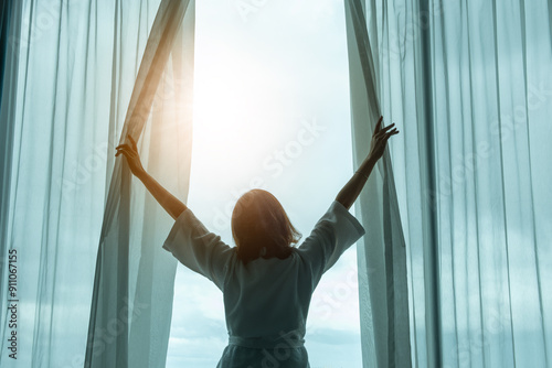The woman was at the window in the bedroom. She opened the curtains on the window. In the morning and she looks at the view of mountains and trees at sunrise.