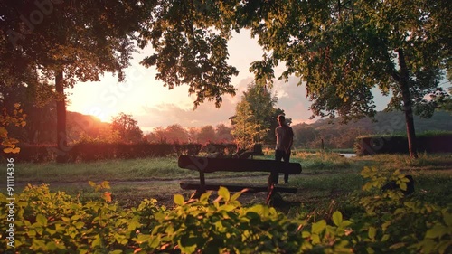 Man walking on a sunlit forest path at sunset finds a bench and sits down to enjoy the gorgeous countryside scenery in an idyllic rural landscape