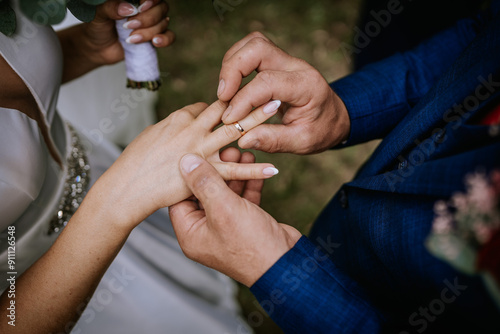 Cesis, Latvia - July 12, 2024 - Close-up of a groom placing a wedding ring on the bride's finger during their ceremony. The bride holds a bouquet in her other hand.