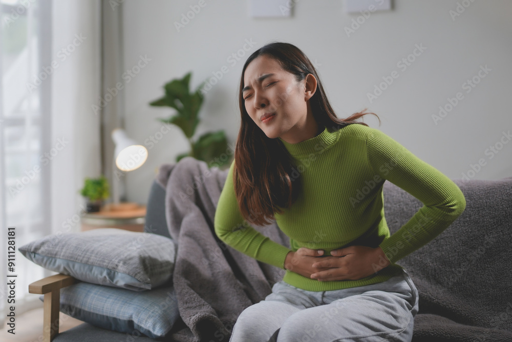 Obraz premium Young woman is suffering from strong stomach pain while sitting on the sofa at home