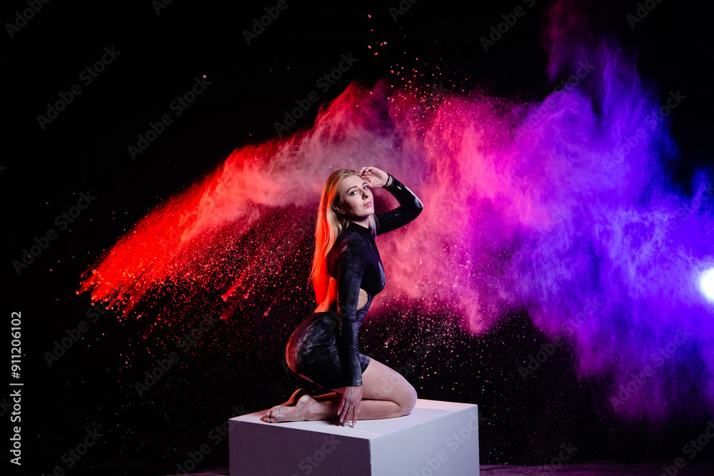 Young slim woman posing on the big white cube with spread multicolored particles in the air