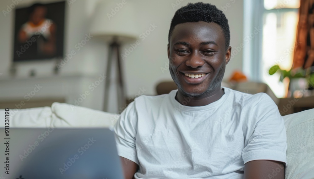 Young Man Smiling While Using Laptop at Home During Daytime in Cozy Living Room Young Man Smiling While Using Laptop at Home During Daytime in Cozy Living Room