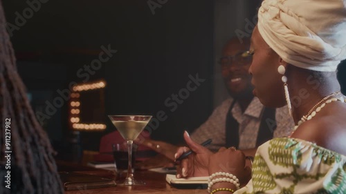 African American woman excitedly talking to her male bandmates while sitting at bar counter with drinks and notepads composing new song together photo