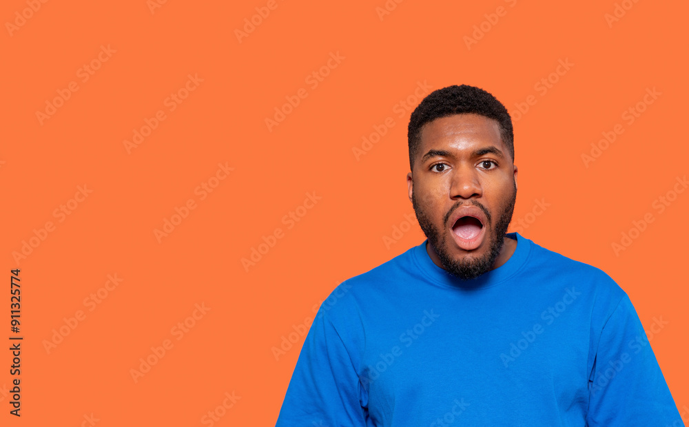 Surprised Young Man Expressing Shock Against Bright Orange Background in Casual Blue Attire Surprised Young Man Expressing Shock Against Bright Orange Background in Casual Blue Attire