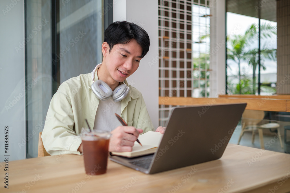 Man is seated at a wooden table in a modern cafe, working with a laptop and writing in a notebook. Man is seated at a wooden table in a modern cafe, working with a laptop and writing in a notebook.