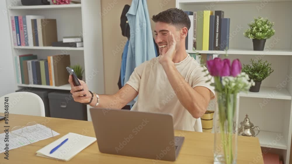 A cheerful hispanic man video calling at his modern apartment with a laptop, phone, flowers, and bookshelf. A cheerful hispanic man video calling at his modern apartment with a laptop, phone, flowers, and bookshelf.