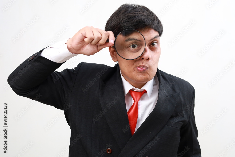 Young asian businessman in formal suit looks into a magnifying glass and makes a funny face Young asian businessman in formal suit looks into a magnifying glass and makes a funny face