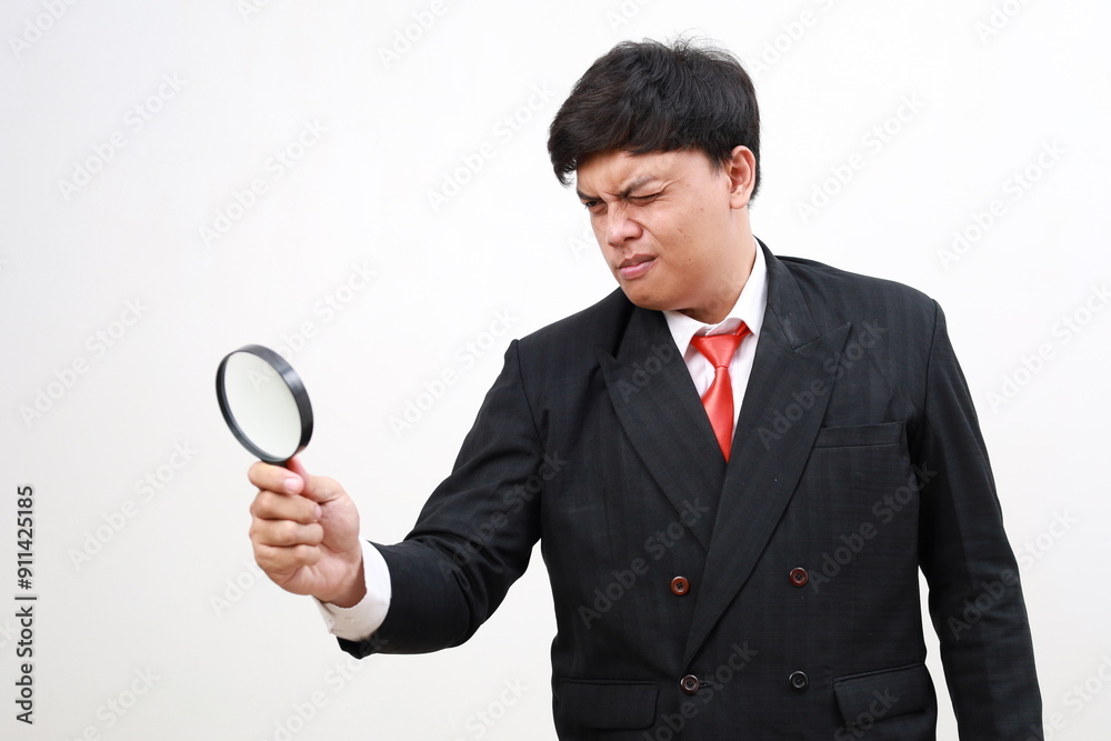 Curious Young asian businessman in formal suit looks into a magnifying glass. Isolated on white Curious Young asian businessman in formal suit looks into a magnifying glass. Isolated on white