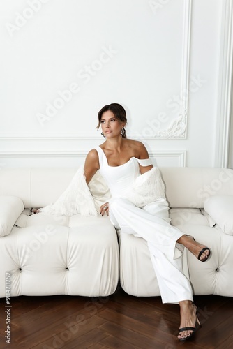 Sophisticated brunette woman in a crisp white pantsuit sits elegantly in a white interior studio. Refined and modern corporate style