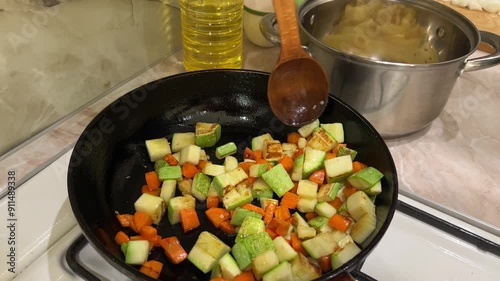 Chef stir frying diced zucchini and carrots in a cast iron skillet on a stovetop with a wooden spoon, preparing a healthy vegetarian meal