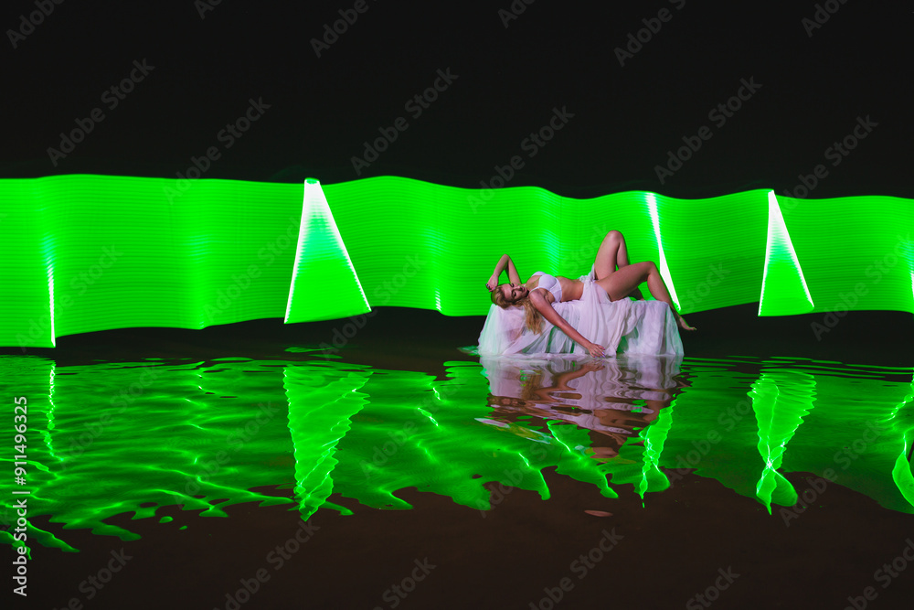 Young slim girl in white swimsuit posing in water at night with abstract green light effect
