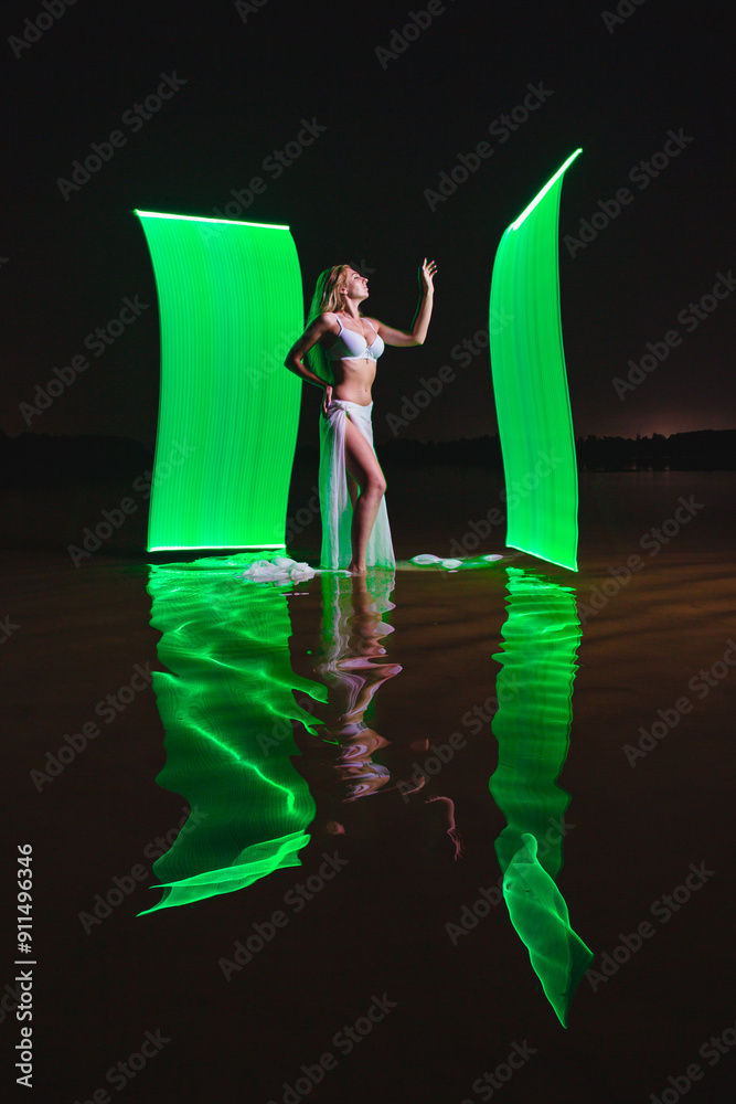 Young slim girl in white swimsuit posing in water at night with abstract green light effect