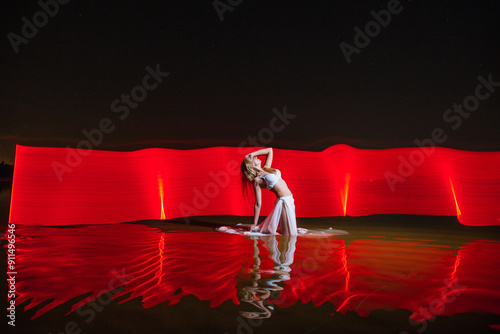Young slim girl in white swimsuit posing in water at night with abstract red light effect