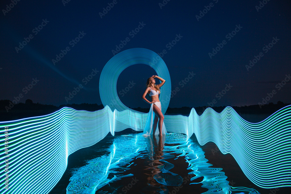 Young slim girl in white swimsuit posing in water at night with abstract blue light effect