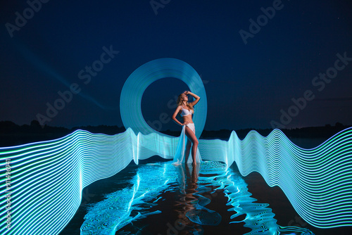 Young slim girl in white swimsuit posing in water at night with abstract blue light effect