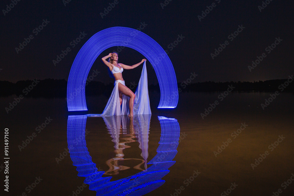 Young slim girl in white swimsuit posing in water at night with abstract blue light effect