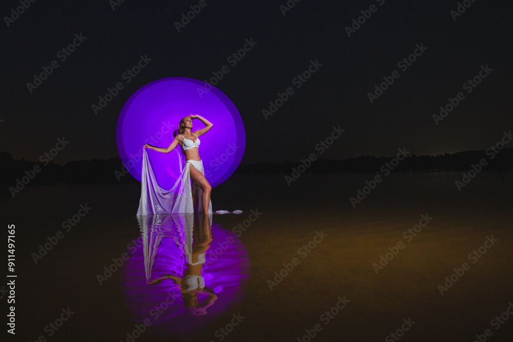 Young slim girl in white swimsuit posing in water at night with abstract purple light effect