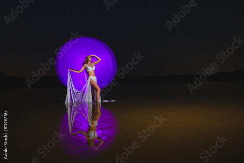 Young slim girl in white swimsuit posing in water at night with abstract purple light effect
