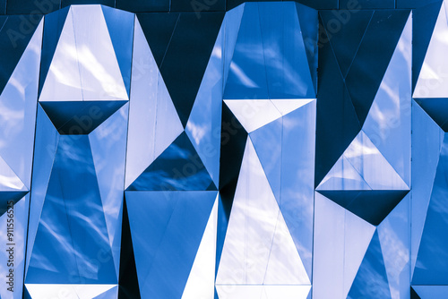 A wall of a building with a geometric pattern composed of multifaceted mirror-like blue panels reflecting light and the sky photo