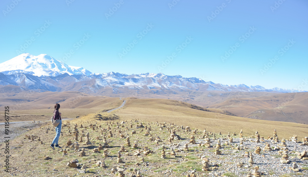 A happy female traveler with a backpack behind her back and her arms raised up rejoices at her trip to the mountains. Elbrus on a clear autumn day. View of Elbrus, North Caucasus, Russia