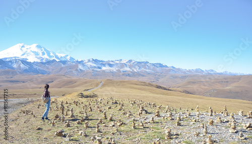 Obraz na płótnie A happy female traveler with a backpack behind her back and her arms raised up rejoices at her trip to the mountains. Elbrus on a clear autumn day. View of Elbrus, North Caucasus, Russia w salonie
