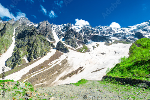 A mountain path among snow and green slopes leading to the rocky snow-covered slope of a high mountain.  The North Caucasus. Russia photo