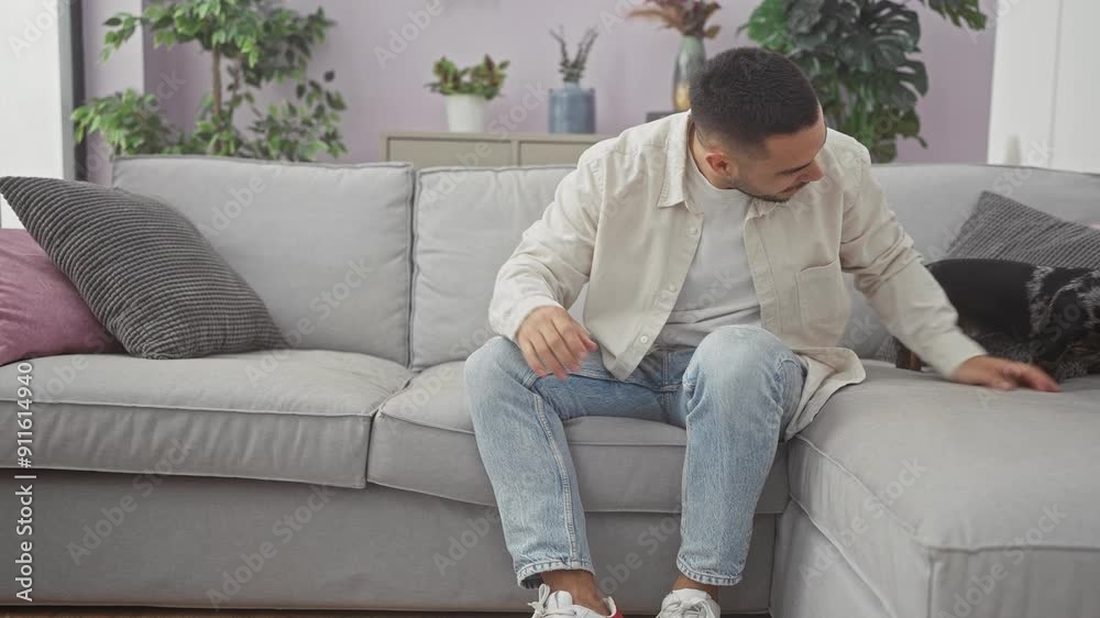 A young hispanic man relaxing at home, affectionately interacting with his pet teckel on a grey couch in a cozy living room. A young hispanic man relaxing at home, affectionately interacting with his pet teckel on a grey couch in a cozy living room.