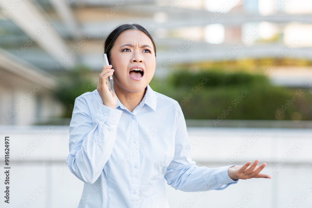 A business professional Asian lady stands outdoors, visibly frustrated while speaking on the phone. She gestures with her hands, indicating her displeasure, copy space A business professional Asian lady stands outdoors, visibly frustrated while speaking on the phone. She gestures with her hands, indicating her displeasure, copy space