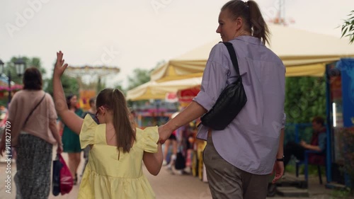 Mother and her young daughter with Down syndrome enter amusement park. Girl in yellow dress points excitedly while holding her mother's hand. Scene highlights parenting child with special needs
