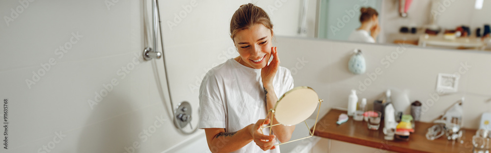 Smiling young woman looking in small round mirror and checking skin while standing in bathroom Smiling young woman looking in small round mirror and checking skin while standing in bathroom