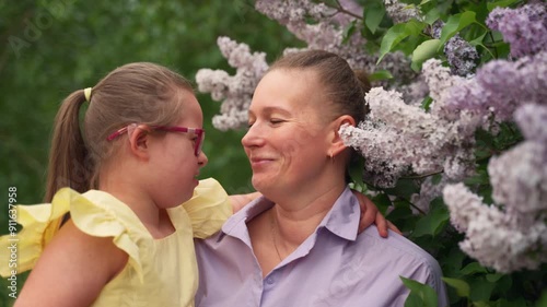 Amidst blooming lilacs, mother and her young daughter with Down syndrome share tender kiss and embrace. Girl is wearing yellow dress, and moment reflects love and support children with special needs