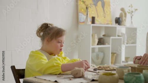 Child with Down syndrome participating in pottery workshop, concentrating on clay art, yellow shirt and apron, white brick wall background. Showcasing social inclusion and acceptance in environments