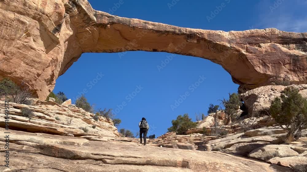 Female hiker walking up a rocky trail towards a natural sandstone formation known as Owachomo Bridge with a bright blue sky behind. Natural Bridges National Monument, Utah, USA. Female hiker walking up a rocky trail towards a natural sandstone formation known as Owachomo Bridge with a bright blue sky behind. Natural Bridges National Monument, Utah, USA.