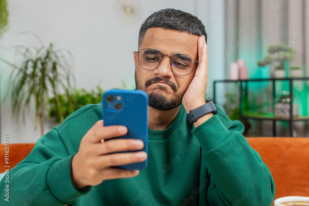 Sad displeased Arabian man use smartphone typing browsing, loses becoming surprised sudden lottery game results, bad news, fortune loss fail, deadline, virus. Young Indian guy at home office at table Sad displeased Arabian man use smartphone typing browsing, loses becoming surprised sudden lottery game results, bad news, fortune loss fail, deadline, virus. Young Indian guy at home office at table