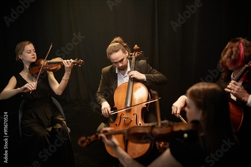 Male classical music artist passionately playing cello with string quartet musicians sitting in circle in studio with contrast top light against black curtains
