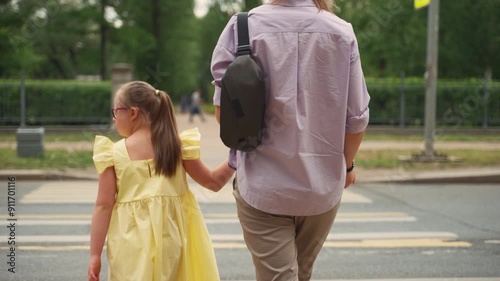 A young girl with Down syndrome and her mother are crossing a street at a crosswalk. The girl, wearing a yellow dress and red glasses, holds her mother's hand. Concept parenting special needs children