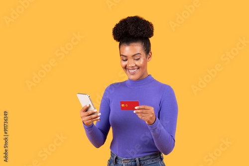 A young woman smiles as she looks at her smartphone while holding a credit card in her other hand. She is wearing a purple long-sleeve shirt and denim jeans