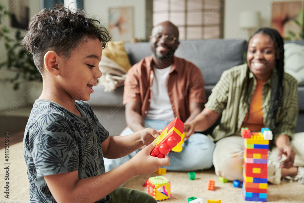 Obraz premium Side view of adorable little boy in casual attire sitting on the floor in front of his parents and playing constructor at leisure