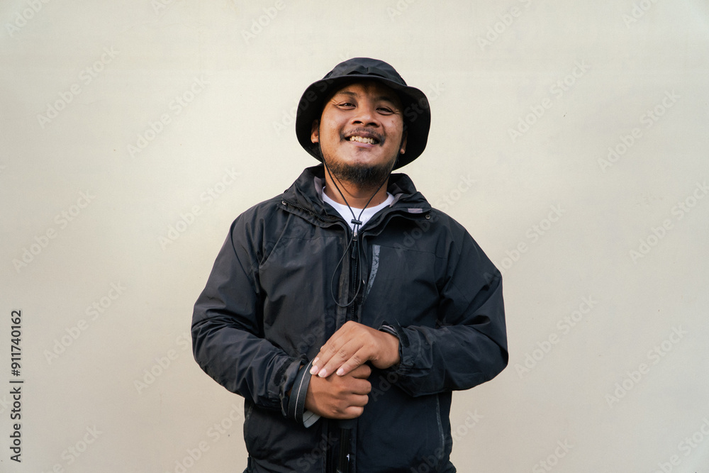 Photo of Asian man wearing all black adventure outfit. Half body portrait of an adult adventurer posing wearing a jacket, boonie hat and trekking poles isolated on a beige background Photo of Asian man wearing all black adventure outfit. Half body portrait of an adult adventurer posing wearing a jacket, boonie hat and trekking poles isolated on a beige background