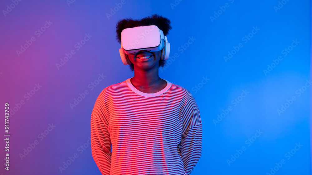 Profile of smiling young African American with VR headset at neon light background exploring metaverse innovation technology through education online learning virtual interface graphic. Contrivance. Profile of smiling young African American with VR headset at neon light background exploring metaverse innovation technology through education online learning virtual interface graphic. Contrivance.
