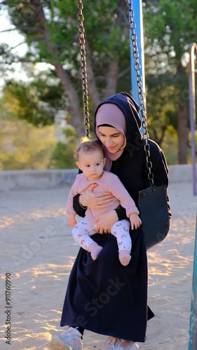 A mother sitting on a swing, holding her baby girl in the park, enjoying the fresh air together.
