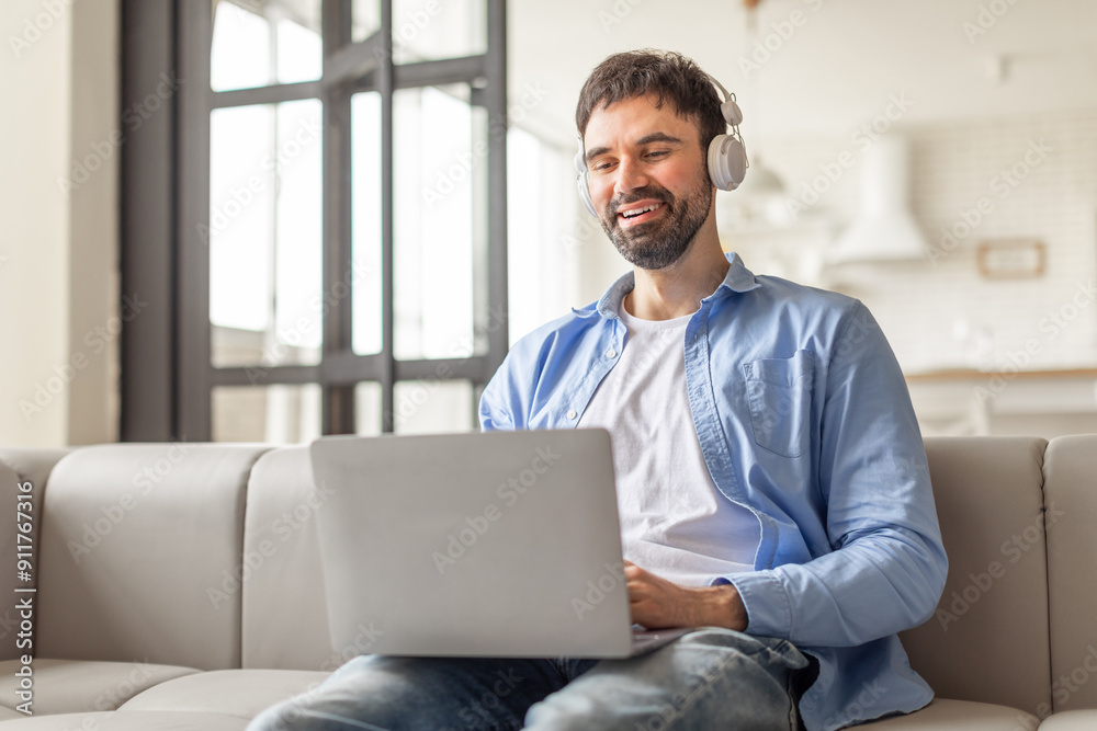 A man sits on a white couch in a modern living room, wearing headphones and smiling while using a laptop. He appears to be enjoying his time online, possibly watching a movie, working on a project. A man sits on a white couch in a modern living room, wearing headphones and smiling while using a laptop. He appears to be enjoying his time online, possibly watching a movie, working on a project.