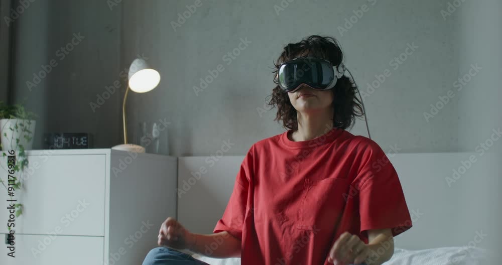 A woman in casual clothes and wearing a virtual reality headset moves her arms while sitting on a bed at home. Daylight