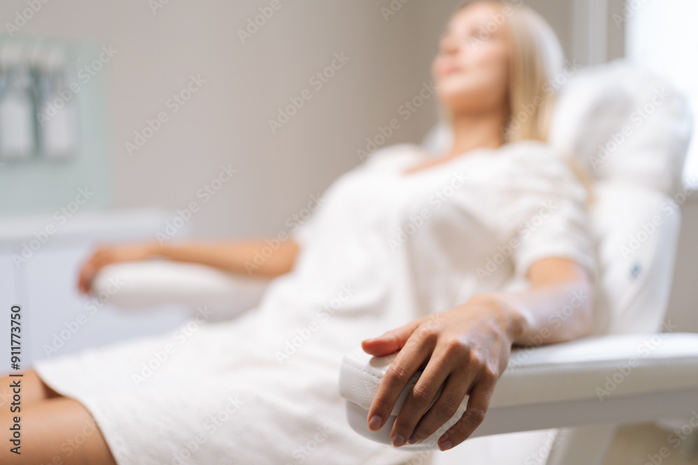 Selective focus shot of blonde young woman client in white procedure uniform lying on cosmetology chair waiting beautician specialist in beauty salon. Concept of cosmetology, spa, beauty treatment.